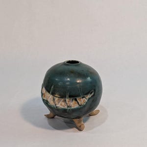 May include: A round, dark teal ceramic vessel with a glossy finish, featuring a wide, horizontal mouth opening lined with pointed, off-white teeth. The object is supported by three small, light-colored, unglazed ceramic feet at the base.
