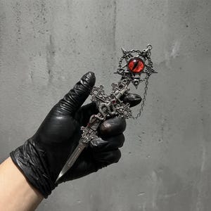 Puede incluir: Una daga de metal plateado con un mango en forma de cruz ornamentada envuelto en un motivo de serpiente. La empuñadura está coronada por un marco circular de pentagrama que contiene un ojo de cristal rojo con una pupila negra vertical.