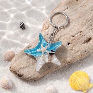 May include: A starfish-shaped keychain made of clear resin, featuring a blue gradient top section and a bottom section filled with small white sand, a tiny beige starfish, and two small spiral seashells attached to a silver metal ring.
