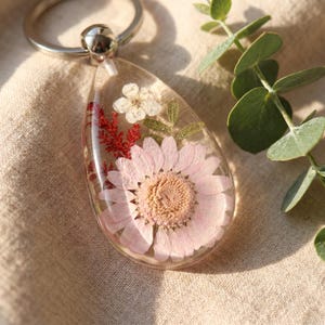 Pode incluir: Um porta-chaves em forma de lágrima feito de resina transparente contendo uma grande margarida rosa claro, uma pequena flor branca e folhagem vermelha semelhante a uma samambaia. O pendente está preso a um anel metálico prateado.