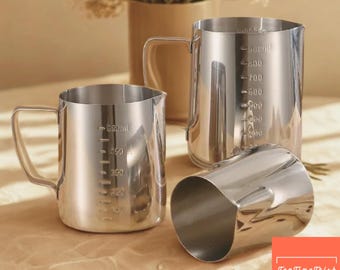 May include: Stainless steel measuring pitchers with handles and measurement markings. The pitchers range in size, with one labeled up to 900ml. The image also includes a small, angled pitcher. The background features a vase with dried flowers.