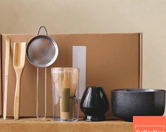 May include: A matcha tea set includes a bamboo whisk, scoop, and spoon, a stainless steel strainer, a black ceramic chawan bowl, and a black ceramic whisk holder. The set is displayed in front of a brown cardboard box.