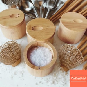 以下が含まれることがあります： 木製の塩入れと、お揃いの蓋と小さなスプーン。 1つの塩入れが開いており、白い塩が見えます。 2つの小さな織りホイップと様々なキッチン用品が背景にあります。 塩入れは明るい色の木材で作られています。