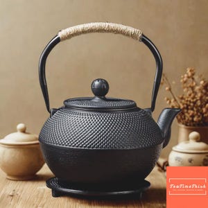 Puede incluir: Tetera de hierro fundido negro con una superficie texturizada y un asa envuelta en cuerda. La tetera tiene un pico y una tapa con un pomo. La tetera se asienta sobre una base negra a juego. La imagen también incluye dos pequeños recipientes de color crema.