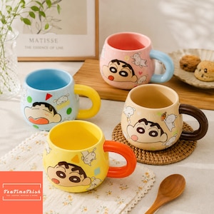 Könnte beinhalten: Vier Keramikbecher mit der Figur Shin-chan. Ein Becher ist hellblau mit gelbem Henkel, einer rosa mit hellblauem Henkel, einer beige mit dunkelbraunem Henkel und einer gelb mit orangefarbenem Henkel. Jeder Becher zeigt verschiedene Cartoon-Grafiken.