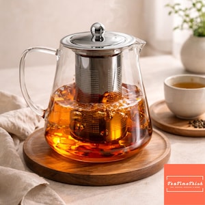 Op de afbeelding: Een glazen theepot met een roestvrijstalen infuser en deksel, gevuld met gezette thee. De theepot staat op een houten dienblad, met een kopje en schotel op de achtergrond. De afbeelding bevat de tekst "TeaTimeTrish".
