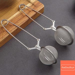 Puede incluir: Dos infusores de té de acero inoxidable con una bola de malla y mango largo. Los infusores están abiertos, listos para ser llenados con hojas de té sueltas. Los mangos tienen un diseño de resorte para facilitar su uso. Fondo de madera y superficie gris.