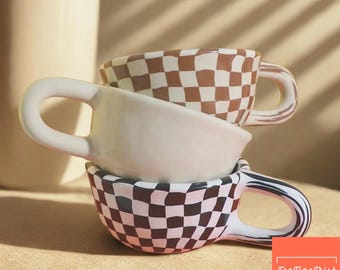 May include: Three ceramic coffee cups stacked on top of each other. The top cup is a solid cream color, the middle cup is a brown and white checkered pattern, and the bottom cup is a black and white checkered pattern. A tall cream-colored mug is in the background.