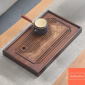 May include: A dark brown wooden tea tray with a carved mountain landscape design and a small black and gold tea pot with a wooden handle. The tray has a rectangular shape with a slotted design.
