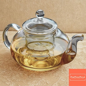 Może przedstawiać: Przezroczysty szklany czajniczek o zaokrąglonym korpusie, zakrzywionym uchwycie i dziobku. Czajniczek zawiera jasno bursztynowy płyn, prawdopodobnie herbatę, i ma szklany kosz do zaparzania w środku. Pokrywka ma mały szklany guz.