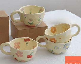 May include: Four ceramic teacups with floral designs. The cups are a pale yellow with various hand-painted flowers in pink, orange, and blue. Each cup has a rounded handle and a wavy rim. One cup is on a cardboard box.