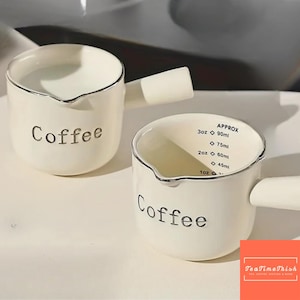 May include: Two white ceramic coffee measuring cups with black text that reads "Coffee". One cup has measurements in ounces and millilitres. Each cup has a handle and a silver rim. The cups are on a white surface.