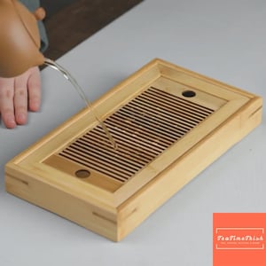 Könnte beinhalten: Ein rechteckiges Teetablett aus Bambus mit einem Lattenentwässerungsbereich. Wasser wird aus einer braunen Teekanne in das Tablett gegossen. Das Tablett hat eine helle Holzoberfläche und ein schlichtes, funktionales Design. Das Logo "TeaTimeThish" befindet sich unten rechts.