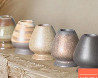 May include: A row of ceramic matcha tea whisks in various colors, including brown, beige, and gray. The whisks are displayed on a decorative wooden surface. The image includes the text "TeaTimeTrish".