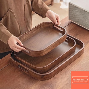 Könnte beinhalten: Drei Holzserviertabletts in verschiedenen Größen, gestapelt auf einer Holzoberfläche. Die Tabletts sind oval mit integrierten Griffen und einer dunkelbraunen Oberfläche. Das Bild enthält den Text "TeaTimeThish".