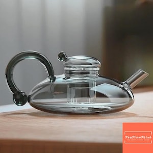 Może przedstawiać: Przezroczysty szklany czajniczek o zaokrąglonym korpusie, zakrzywionym uchwycie i dziobku. Czajniczek ma pokrywkę z małym pokrętłem i wewnętrznym zaparzaczem. Czajniczek stoi na drewnianej powierzchni.