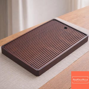 Op de afbeelding: Een rechthoekige, donkerbruine houten theeblad met een lattenoppervlak en een klein gaatje. De lade staat op een lichtgekleurde placemat en een houten tafel. De lade is ontworpen voor de bereiding van thee.