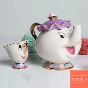 May include: A white ceramic teapot and teacup set with cartoon-like facial features. The teapot has a purple lid and gold handle. The teacup has a gold handle and a pink and blue base. The set is inspired by a popular animated film.