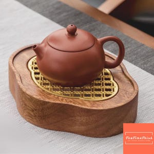 Puede incluir: Una pequeña tetera de cerámica marrón con tapa a juego descansa sobre una bandeja de madera con una rejilla de metal dorada. La bandeja es de color marrón claro y tiene forma curva. La tetera tiene un cuerpo redondeado y un asa.
