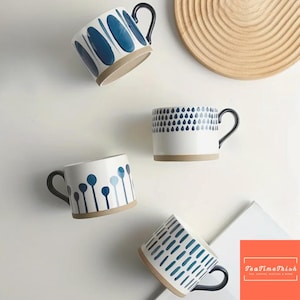 Könnte beinhalten: Vier weiße Keramikbecher mit blauen Mustern und schwarzen Griffen werden gezeigt. Die Becher haben unterschiedliche Designs, darunter ovale Formen, Tropfen und vertikale Linien. Die Becher haben einen beigefarbenen Boden. Im Hintergrund befindet sich ein Holztablett. Das Logo "TeaTimeTrish" befindet sich unten rechts.