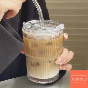 Op de afbeelding: Een heldere, geribbelde glazen beker gevuld met ijskoffie, met een doorzichtig deksel en rietje. De drank heeft een gelaagde uitstraling met een lichtbruine kleur. Het glas wordt door een persoon vastgehouden. De rechter benedenhoek heeft een logo met "TeaTimeThish".
