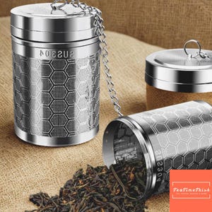 Può includere: Infusore per tè in acciaio inossidabile con catena e coperchio. L'infusore ha un motivo a nido d'ape e il testo "202304" sul lato. Foglie di tè sfuse si rovesciano. Sfondo neutro.