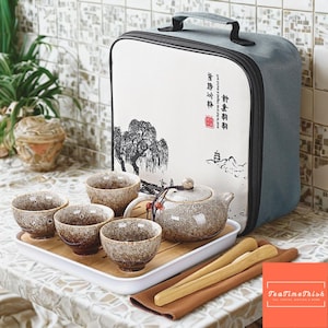 Pode incluir: Um conjunto de chá de cerâmica com uma chaleira, quatro xícaras e utensílios de bambu em uma bandeja branca. O conjunto tem um acabamento marrom e bege salpicado. Uma mala de transporte com um design de paisagem e texto está atrás do conjunto de chá.