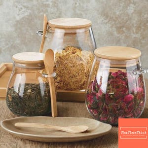 Può includere: Barattoli di vetro trasparente con coperchi e cucchiai di legno, riempiti di erbe e fiori secchi. Un piccolo piatto di ceramica contiene un cucchiaio. L'immagine include il testo "TeaTimeTrish".