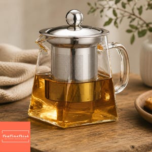 May include: A clear glass teapot with a stainless steel infuser and lid. The teapot is filled with amber-colored tea. The handle is curved, and the teapot sits on a wooden surface. The logo "TeaTimeTrish" is in the bottom left corner.