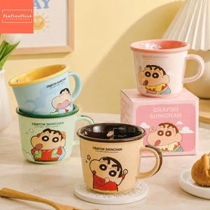 Könnte beinhalten: Drei Keramikbecher mit Crayon Shinchan-Figuren in Hellblau, Hellgrün und Beige. Jeder Becher hat einen Henkel, einen Rand und den Schriftzug Crayon Shinchan. Ein beiger Becher steht auf einem weißen Untersetzer, dahinter eine rosa Schachtel.
