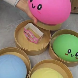 May include: A set of four round, smooth, squishy toys shaped like steamed buns, each in a different color: pink, green, blue, and yellow. Each bun features a printed face with black eyes, a smile, and pink cheeks. They are housed in individual round bamboo-style containers, with one small rectangular paper box labeled STEAMED BUN included.