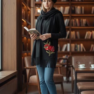 May include: A woman wearing a black beret with a red floral design, a black scarf with a red floral embroidery, a black long-sleeve shirt, and blue jeans. She is holding a book in a library setting.