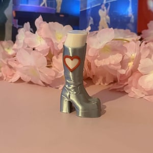 以下が含まれることがあります： ブロックヒールとプラットフォームソールを備えたシルバーカラーのミニチュアブーツ。ブーツの側面には、赤い縁取りの白いハートのエンブレムがあしらわれています。ブーツの上部は開いており、滑らかで明るい色の内面が見えています。