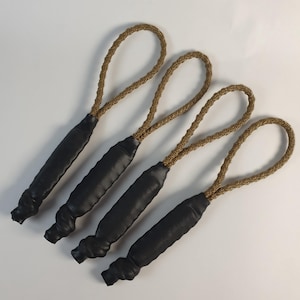 May include: Four black leather zipper pulls with braided tan cords. The pulls have a rounded, textured design at the base and a loop at the top. The items are arranged diagonally against a white background.