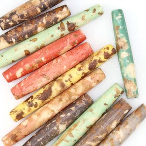 Puede incluir: Un surtido de cuentas cilíndricas de colores en tonos marrón, verde, amarillo y rojo, con un aspecto moteado y envejecido. También se ven una geoda, un cristal y un collar de cuentas de madera.