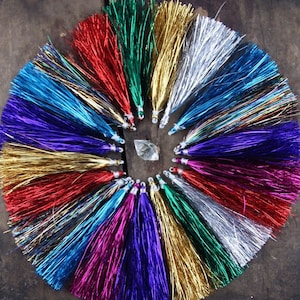 May include: A collection of colorful metallic tassels arranged in a circle on a wooden surface. The tassels are made of shiny, iridescent material and come in a variety of colors, including red, gold, silver, blue, green, and purple.
