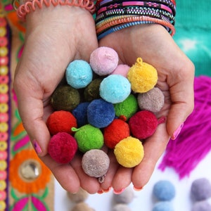 May include: A collection of colorful pom poms in various shades of red, green, blue, yellow, and pink. The pom poms are held in a person's hands.