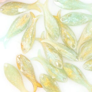 May include: A collection of pale green and gold fish-shaped beads scattered on a white surface. A tassel, a strand of gold beads, and a colorful crystal are also present. The beads are likely for jewelry making.