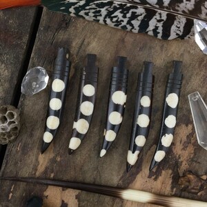 May include: Six black and white spotted bone-like pendants with a rounded tip. The pendants are arranged in a row on a wooden surface.