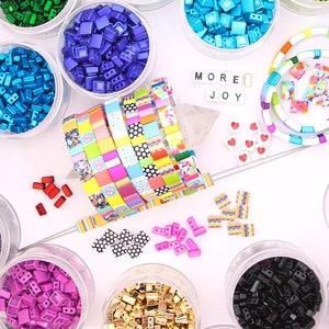 May include: A selection of colourful beads in clear plastic pots. The beads are in a variety of colours, including red, yellow, green, blue, and purple. A woven bracelet and the words "MORE JOY" are also visible.