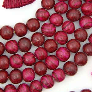 Puede incluir: Una hebra de cuentas de madera de color rosa oscuro con un patrón de remolino. Las cuentas están enhebradas en un cordón dorado y tienen aproximadamente 1 cm de diámetro. Las cuentas se muestran con dos borlas, una rosa y otra rosa oscuro.