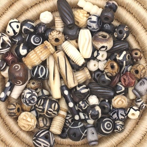 May include: A collection of black, white, and brown bone beads in a woven basket. The beads are of various shapes and sizes, including round, cylindrical, and oval.