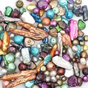 May include: A collection of colorful, irregularly shaped pearls in various sizes and hues. The pearls include shades of pink, purple, green, blue, and gold. Some pearls have a lustrous sheen, while others have a more matte finish. The pearls are scattered on a white surface.