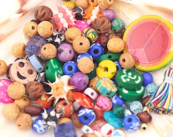May include: A collection of colorful beads in various shapes and sizes, including wooden, glass, and plastic beads, displayed in a seashell. The beads are in shades of pink, blue, green, yellow, and brown. A pink sticker with the text "Little beads, Big dreams" is also visible.
