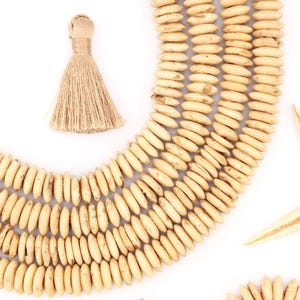 以下が含まれることがあります： 白い背景に、自然な色合いの木製ビーズネックレス、タッセル、貝殻、ヒトデのコレクション。ビーズは楕円形で、曲線に配置されています。タッセルはベージュ色の繊維でできています。