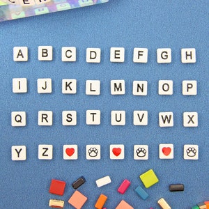 May include: A colorful beaded bracelet spelling "CENTER" rests on a clear crystal. Individual letter tiles and additional colorful beads are scattered on a blue surface. Alphabet tiles are arranged in rows, with heart and paw print tiles.