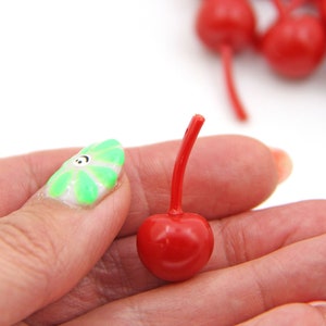May include: A single red cherry with a green stem, held in a hand with a green and white nail design. The cherry is a small, round fruit with a glossy finish.