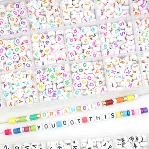 May include: A white plastic storage box filled with colourful letter beads. The beads are arranged in rows and columns, with each row containing a different letter of the alphabet. The box also includes two bracelets with colourful beads that spell out the phrases "DREAM BIG" and "YOU GOT THIS".