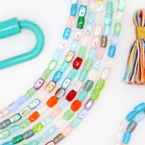 May include: Multiple strands of rectangular, iridescent glass beads in various colors including blue, green, red, orange, and clear. A turquoise carabiner clasp is on the left, and a multicolored tassel with gold thread is on the right.