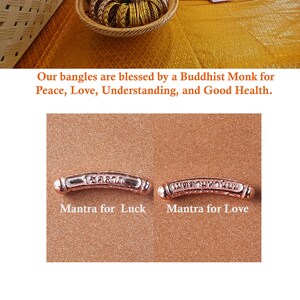 May include: A close-up of two gold-colored metal bangles with engraved Thai script. The bangle on the left is labeled "Mantra for Luck" and the bangle on the right is labeled "Mantra for Love".